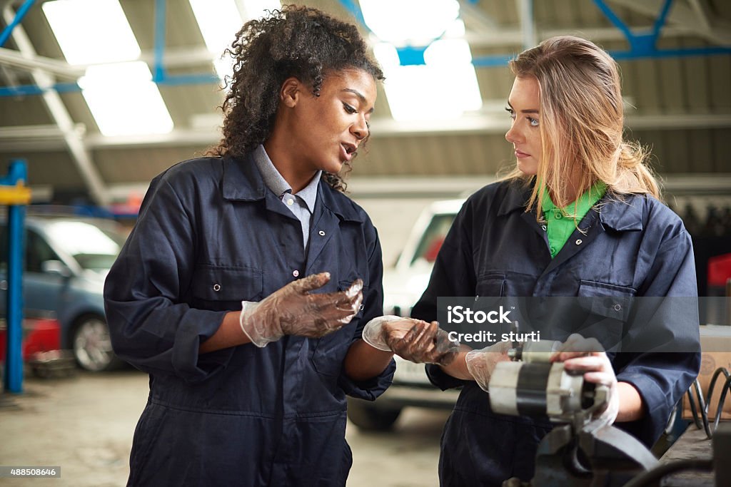 car shop teaching A female mechanic is filing the bushes on an alternator at a work bench in a garage repair shop. She is wearing blue overalls and being advised by a colleague or her boss. They are both female . 20-29 Years Stock Photo