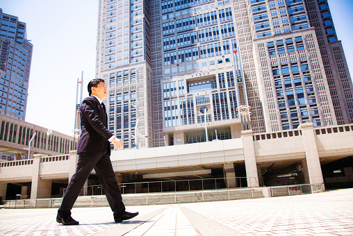 Japanese government worker walking to work in front of Tokyo city hall. Tilted profile view, full length. ei is dressed in a suit and is looking purposefully ahead. Photographed in Shinjuku, Tokyo, Japan.