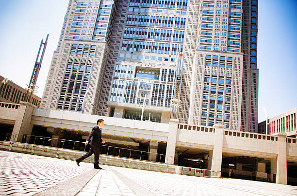 Japanese civil servant working to work at Tokyo City Hall. Tilted profile view, full length. ei is dressed in a suit and is looking purposefully ahead. Photographed in Shinjuku, Tokyo, Japan.