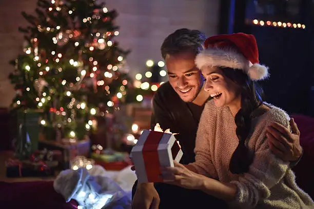 portrait of a young couple in their living room in  christmas eve the young woman with santa hat on head smiled in opening his gift, a lit Christmas tree in the background