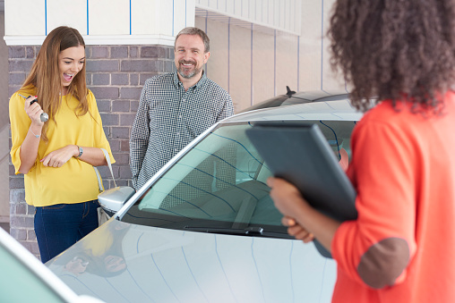 A mature man and his young daughter are looking at  cars in a car showrooms . A saleswoman is explaining about the car whilst the young woman clutches the keys to her new car and screams with delight.