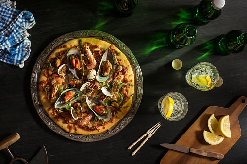 High resolution, full frame, digital capture of a thin crust pizza topped with various shellfish including clams, shrimp, and mussels; mozzarella, green peppers, rosemary, and fresh chiffonade basil leaves. Bottles and glasses of sparkling mineral water, lemons, cutting board and knife, seafood picks, kitchen towel, and a pizza cutter are inset.