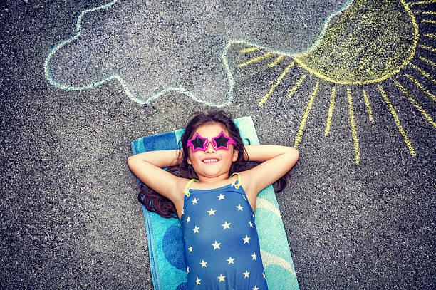 Happy little girl outdoors Happy little girl wearing swimsuit and stylish sunglasses lying down on the asphalt near picture of the sun comes out from behind the clouds, cute baby needs of summer holidays summer camp stock pictures, royalty-free photos & images