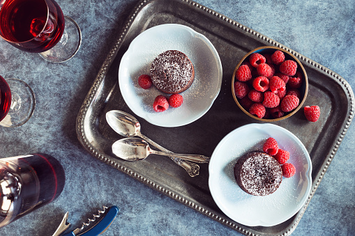 A metal tray holds two porcelain plates with baked chocolate lava cakes topped with powdered sugar and raspberries. A bowl of fresh berries sits next to the two desserts. A bottle of red wine with two glasses and a corkscrew is at the opposite end of the blue table.