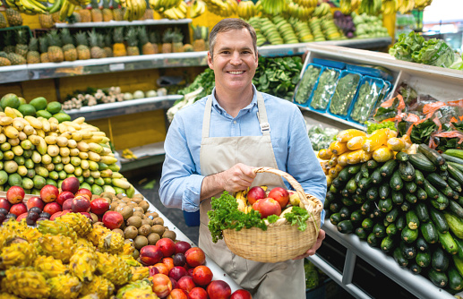 Happy man working at the food market and carrying a basket of fruits and vegetables while looking at the camera smiling