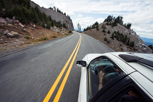 Road trip image of a white car traveling along a mountain pass road in the back country of Oregon's rural wilderness. This image shows the driver's hands on the steering wheel, and has intentional motion blur from the movement and speed of the car. Road is a single lane, two way paved road with a turn ahead.
