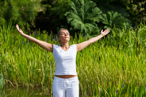 outdoors meditation - gorgeous young yoga woman outstretching arms,closing eyes to focus on inner beauty to relax and meditate with green bushy background and summer daylight