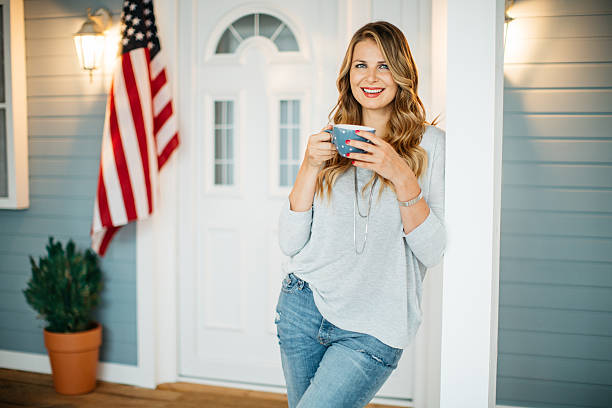 Woman in front of a house. Woman standing on a porch of her house, American flag in background. Holding coffee mug. in front of photos stock pictures, royalty-free photos & images