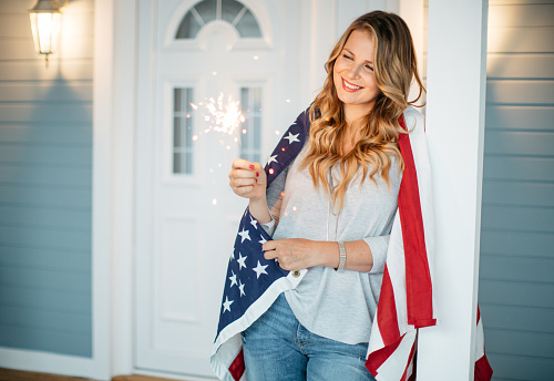 Woman standing on a porch of her house, American flag is on her shoulders. Holding sparkler.