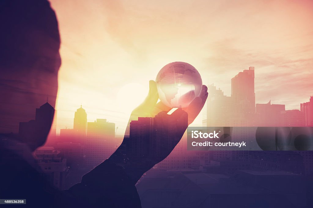 Business man in suit with cityscape montage. Business man in suit with cityscape montage. The man is unrecognizable and you cannot see his face. He is superimposed onto a city skyline at sunset. He is holding a world map globe like a crystal ball. Success, vision concept with copy space. The Way Forward Stock Photo