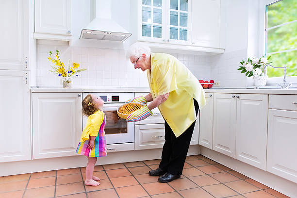 Cute grandmother and little girl baking pie in white kitchen Happy beautiful great grandmother and her adorable granddaughter, cute curly toddler girl in a colorful dress, baking an apple pie together standing next to a white oven in a sunny modern kitchen with big window fume cupboard stock pictures, royalty-free photos & images