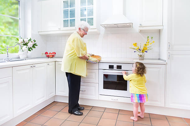 Grandmother and little girl baking in kitchen with big window Happy beautiful great grandmother and her adorable granddaughter, curly toddler girl in colorful dress, baking an apple pie together standing next to white oven in sunny modern kitchen with big window fume cupboard stock pictures, royalty-free photos & images
