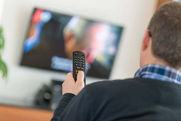 Man using TV Remote Control Man holding a Television remote control, changing channels, selective focus on the foreground with TV on in the background. cable tv stock pictures, royalty-free photos & images