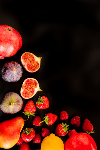 Strawberries, figs, a pear, 2 pomegranates and a lemon, taken overhead on a black background. Space available for text.