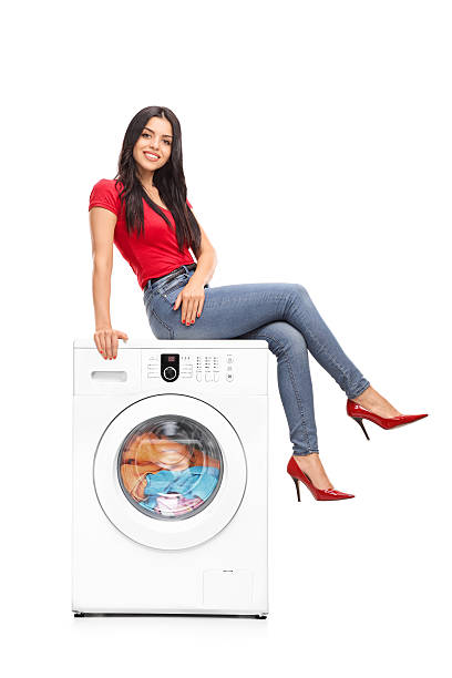 Full length portrait of a beautiful young woman sitting on a washing machine and looking at the camera isolated on white background