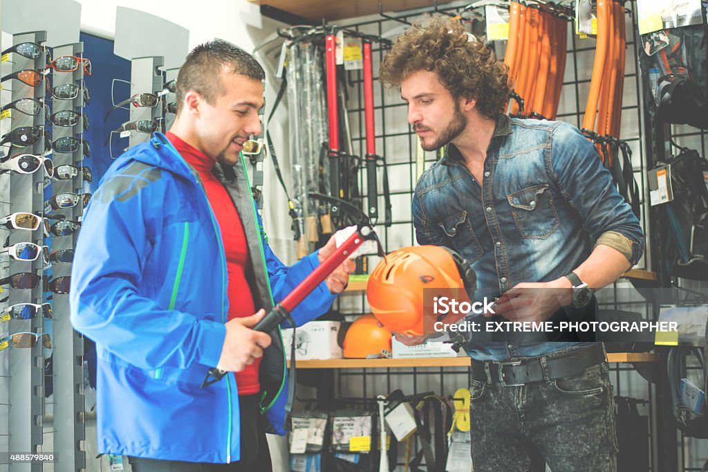 Man buying ice axe in sports mega store Man in sports and outdoor equipment store looking to buy alpine equipment. He is holding the ice axe and the store clerk is holding protective helmet. 2015 Stock Photo