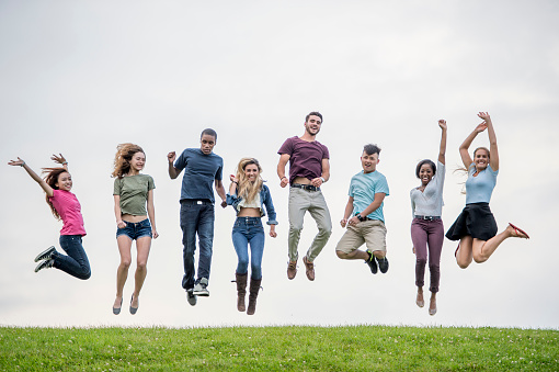 A multi-ethnic group of high school friends are on top of a grassy hill, they have jumped up for the picture and are in mid air, they are smiling and looking at the camera.