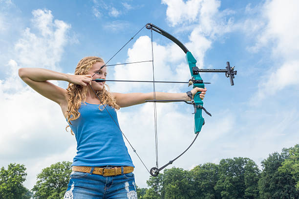 Blonde caucasian girl shooting with arrow and compound bow outdoors on sunny day. The young european woman is aiming at the target in rural area. The dutch teenage girl is tensioning the compound bow and holding the arrow. Don't try this at home because it is a very dangerous sport, high risk. The bow is like a weapon and you are taking a risk. Better have a life insurance. Concept of shot, score, success, win, target, aim, fear, fearful, powerful, cool, sturdy.