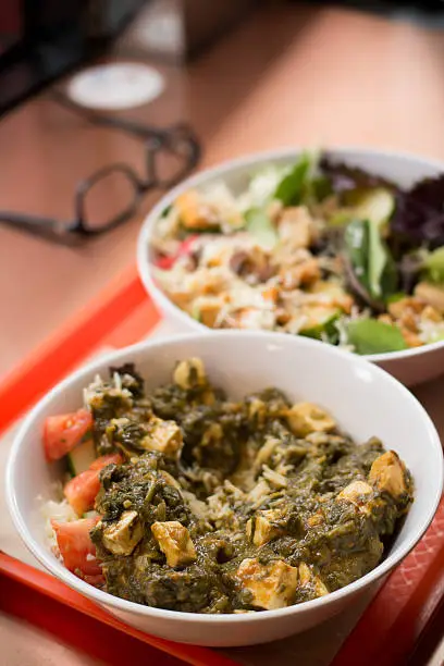 Delicious paneer cheese saag and spinach curry bowl and a  vegan roasted vegetable salad on an orange serving tray. Reading glasses are in the background.  