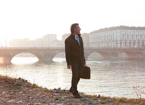 Businessman walks beside river, sunrise, Turin, Piedmont, Italy