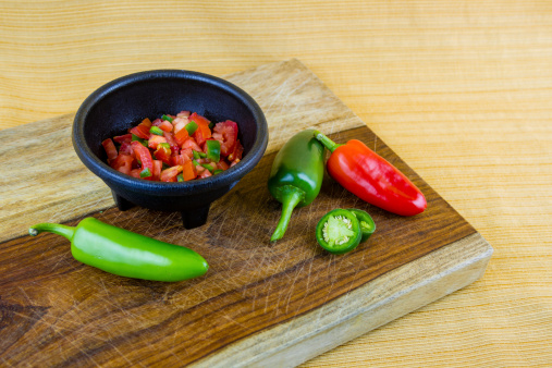 Homemade salsa in bowl with jalapeño peppers,  diced tomatoes, red bell peppers on rustic cutting board
