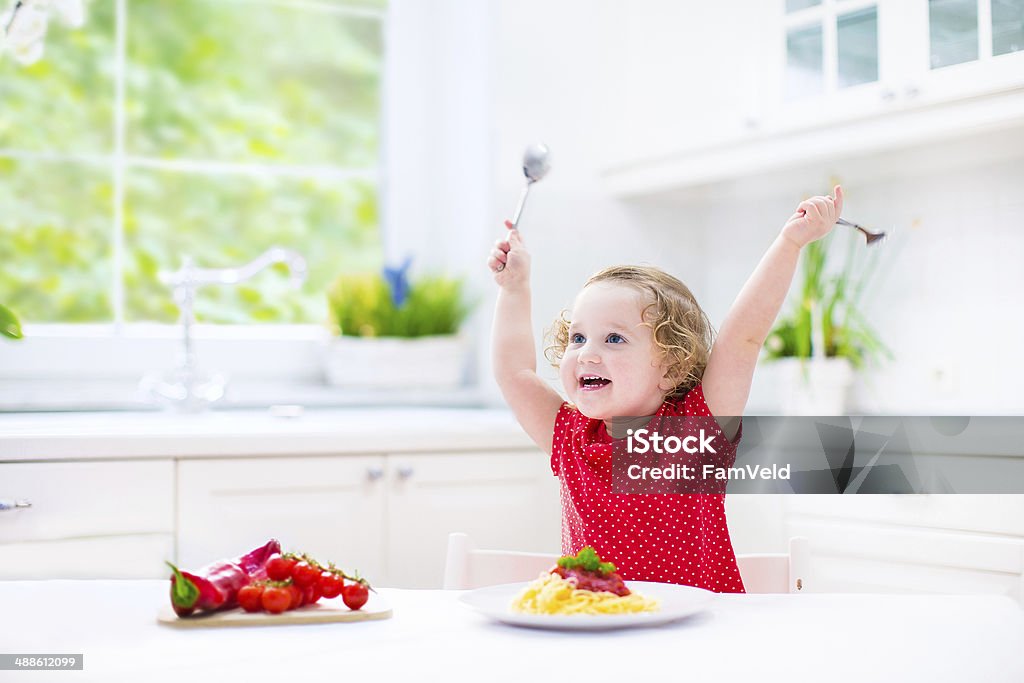 Cute laughing toddler girl with fork and spoon eating spaghetti Cute curly laughing toddler girl in a red shirt playing with fork and spoon eating spaghetti with tomato sauce and vegetables for healthy lunch sitting in a white sunny modern kitchen with big window Baby - Human Age Stock Photo