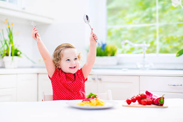 istock Laughing toddler girl playing with fork and spoon eating spaghetti 488612123