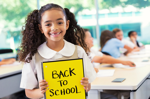 Adorable mixed race African American and Caucasian elementary age schoolgirl is holding a yellow spiral notebook with BACK TO SCHOOL written in black marker. Girl has curly pigtails and is missing her two front teeth. Students are wearing private school uniforms.