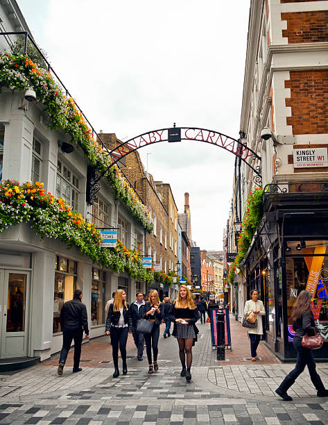 London, England - September 8, 2015: Three beautiful young blonde women walk amongst tourists and shoppers in Carnaby Street, Central London. Carnaby Street became famous in the 1960s for its large number of shops stocking Mod, secondhand and hippie clothing. It is one of the most well known streets visited by tourists to London. Colourful flower displays brighten a cloudy day.