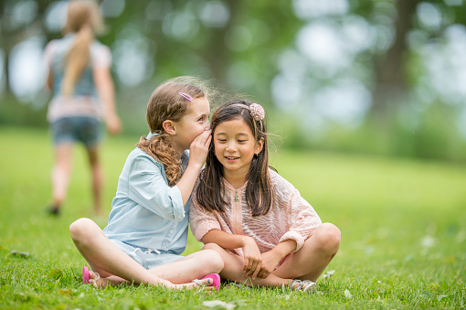 Two little girls are sitting outside in the grass together, one girl is whispering a secret into the other girl's ear.