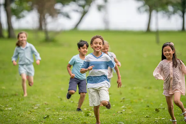 A multi-ethnic group of elementary age children are running together through a park and are playing tag. One little boy is smiling and looking at the camera.