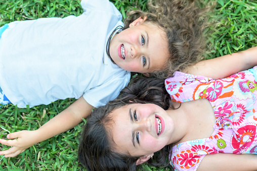 Posing together headshot of a 6 years old little girl with her 3 years old sister lying on the grass smiling from above