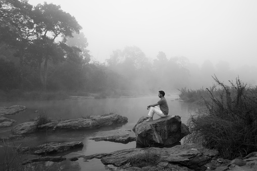 a young man  staring in front of him while sitting on a rock surrounded by water