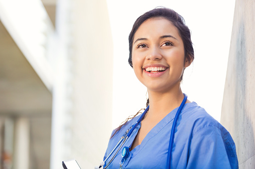 Young adult Hispanic woman is smiling while standing outside on college campus. She is nursing or medical student. Woman is smiling and is wearing blue hospital scrubs. She is also wearing a stethoscope and school ID.