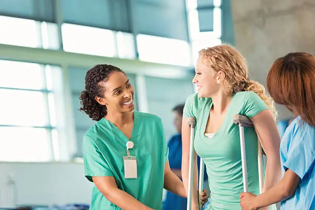 Young adult African American female nurse or physical therapist is helping teenage patient. Teenage Caucasian female patient with blonde curly hair is using crutches and talking to healthcare professional during physical therapy.