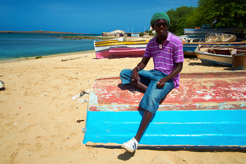 Sal Rei Cape Verde - July 29, 2012: A sunny day in Sal Rei, and a smiling young local sits on an upturned boat on the main beach of Sal Rei, the main settlement of Boa Vista, one of the islands that make up the African nation of Cape Verde. Behind him are various other small brightly coloured boats.