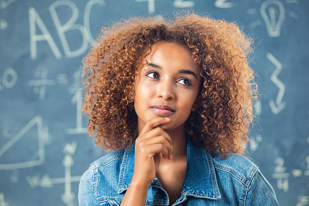 Pretty African American teenage girl in front of blackboard Teenage African American girl is high school student. She is touching her chin while making a thoughtful or puzzled face. High school student is wearing a denim jacket and has curly natural hair. She is standing in front of blackboard or chalkboard, with math symbols written in chalk. ethiopian ethnicity photos stock pictures, royalty-free photos & images