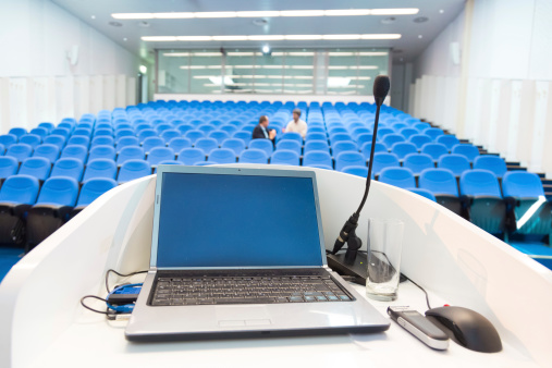 Laptop and microphone on the rostrum in conference hall with blue velvet chairs.