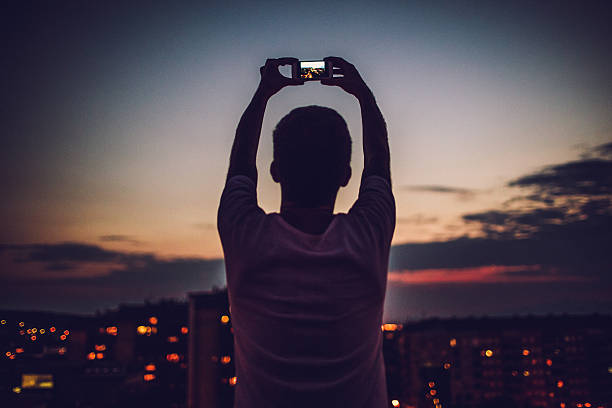 Photographing that perfect time of the day Young man is holding his mobile phone up above his head and capturing that perfect time of the day when sun is going down  street light photos stock pictures, royalty-free photos & images