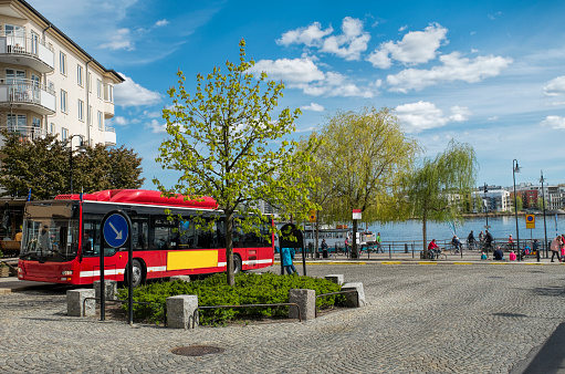 Spring in Stockholm, Sweden. A red bus passes on the street by the Hammarby csnal. . Down by the water edge people are boarding the ferry going over to Hammarby sjöstad one of the newest parts on Stockholm The trees are green and the sky blue with fluffy clouds.