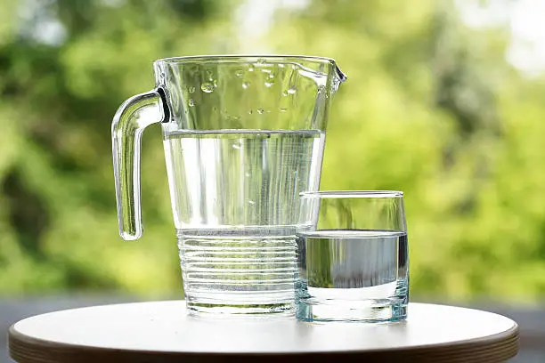 Glass of water near jug on green nature background