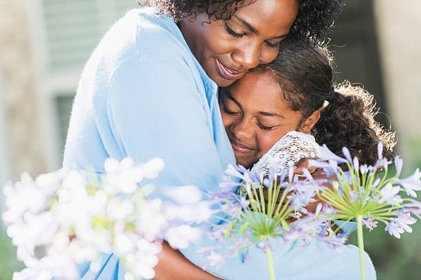 An African American mother and daughter standing together outdoors on a sunny day.  Mom is hugging her little girl, 10 years old, flowers in the foreground.