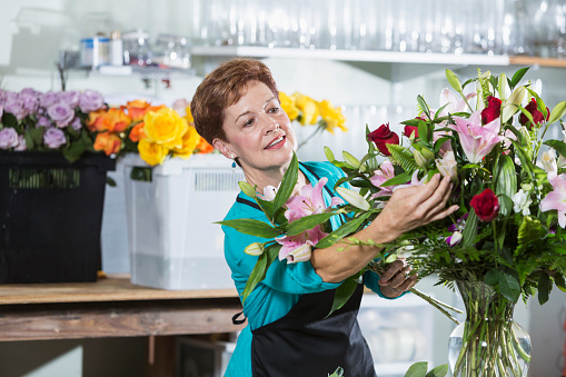 A mature woman in her 60s working as a florist in a flower shop.  She is wearing an apron, arranging flowers in a vase. There are bins full of flowers and assorted glass vases on the table and shelves behind her.