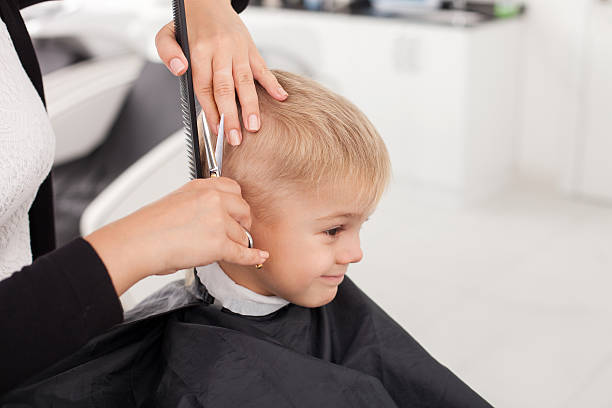 Skilled young female hairstylist is cutting human hair Close up of hands of hairdresser. The woman is standing and making haircut for small boy. She is holding a comb and scissors. The child is sitting and smiling hairstyle stock pictures, royalty-free photos & images