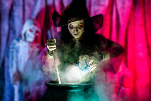 Witch wearing black clothing and hat holds a stick as she prepares her special brew in a cauldron for Halloween evening.  Zombie and tombstone in the background. Purple and pink backdrop. Spooky scene.  Steamy, smokey pot. 