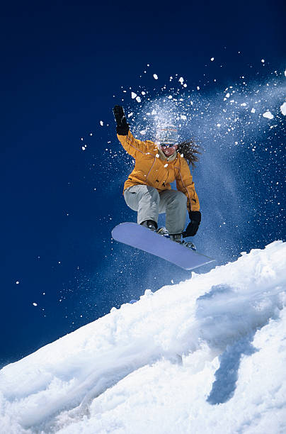 Cheerful young woman having fun in winter mountain, jumping and dancing. Behind friends running in the snow. All with ski wear, goggles, gloves and hats. Snowflakes in the air. As background snow mountain and trees.