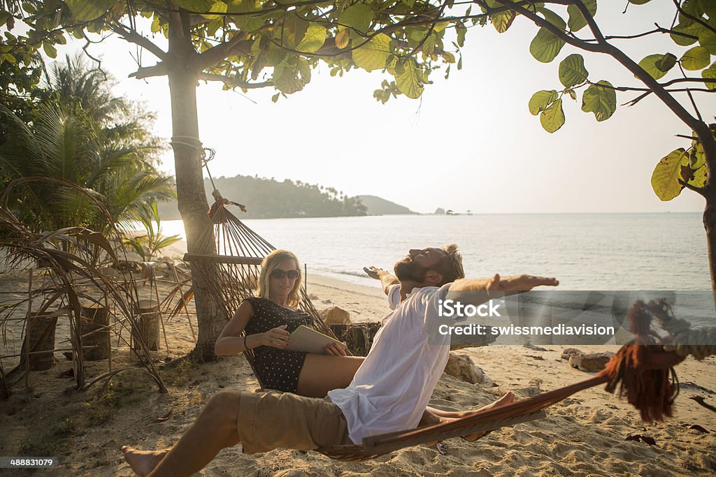 Young couple on hammock relaxing and using digital tablet Young happy couple in holidays enjoying the tropical climate on the Island, relaxing with books and digital tablet on a hammock. Adult Stock Photo