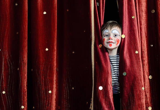 Boy Clown Peering Through Stage Curtains Young Boy Wearing Clown Make Up Peering Out Through Opening in Red Stage Curtains theatrical performance stock pictures, royalty-free photos & images