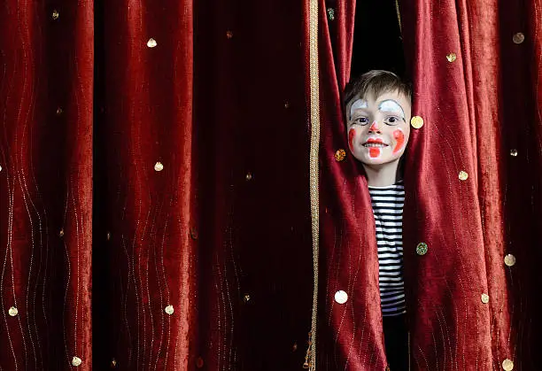 Young Boy Wearing Clown Make Up Peering Out Through Opening in Red Stage Curtains