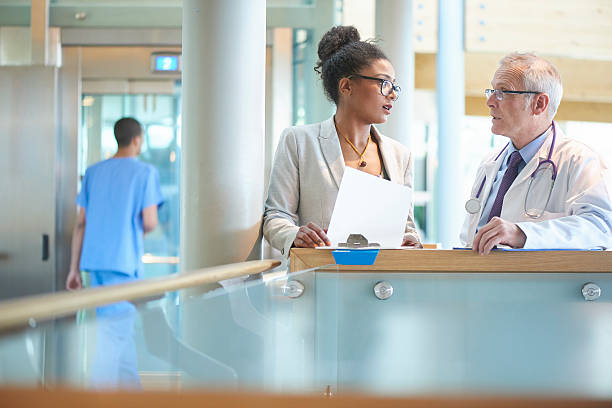 business and medical realtionship. A suited woman and a male doctor wearing a lab coat stand on a stairwell of a modern hospital and discuss some case notes. The woman could be an administrator or business woman . medical office lobby stock pictures, royalty-free photos & images